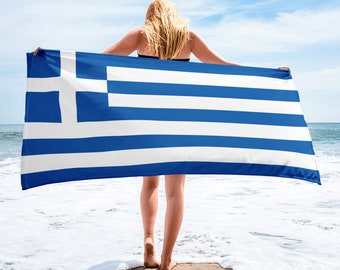 May include: A woman standing on a beach holding a blue and white striped beach towel with the Greek flag design.