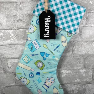 May include: A Christmas stocking with a teal and white gingham cuff and a teal background patterned with baking tools. A black tag with the name "Henry" is attached. The stocking is hanging against a gray brick wall.