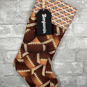 May include: A Christmas stocking with a football theme. The stocking is brown with a pattern of footballs. The cuff is patterned with orange, black, and white diamonds. A black tag with the name "Benjamin" hangs from the stocking.