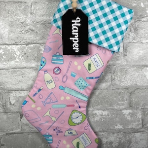 May include: A pink Christmas stocking with a teal and white gingham cuff. The stocking is decorated with baking-themed illustrations and the name "Harper" on a black tag.