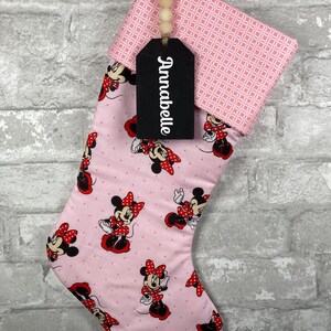 May include: Pink Christmas stocking featuring Minnie Mouse illustrations in red and black. The cuff has a pink geometric pattern. A black tag with the name "Annabelle" hangs from the top, attached with a wooden bead string. The stocking hangs against a gray brick wall.