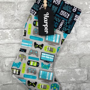 May include: A gray Christmas stocking with a pattern of video game controllers, consoles, and text. The cuff is black with white text. A black tag with the name "Harper" hangs from the top, adorned with wooden beads.