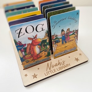 May include: A wooden book display stand holding children's books. The stand is engraved with "Noah's LITTLE LIBRARY" and star cutouts. Two books are visible, one titled "ZOG" and the other "The Scarecrows' Wedding".