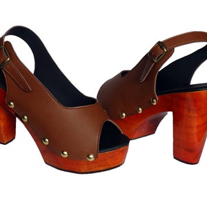 Puede incluir: Un par de sandalias de tacón de cuero marrón con puntera abierta y correa de tobillo ajustable. Los tacones son de madera y tienen un acabado marrón rojizo.