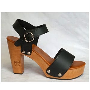 Puede incluir: Sandalias de tacón de cuero negro con plataforma de madera y correa de tobillo con hebilla. Las sandalias tienen tachuelas plateadas en la plataforma y el tacón.