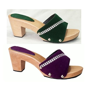 Peut inclure: Deux paires de sabots en bois à talons hauts. L'une présente une bride en daim vert avec des ornements argentés, l'autre une bride en daim violet. Les deux ont une base et un talon en bois naturel.