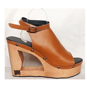 May include: Tan leather wedge sandals with a wooden platform and an ankle strap with a buckle. The platform has a cutout design and is decorated with metal studs.