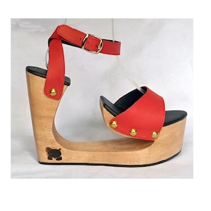 May include: Red leather platform sandals with a wooden wedge heel. The heel is shaped like a curved arch and has a black cat design on the bottom. The sandals have a gold buckle ankle strap and gold studs on the strap and platform.