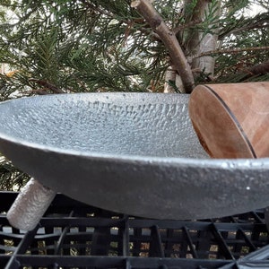 May include: A silver-colored, textured metal bowl with three short legs. A wooden pestle rests inside the bowl. The bowl is set against a backdrop of green foliage.
