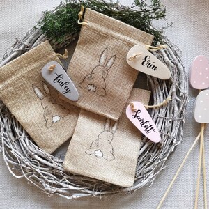 May include: Four small burlap bags with bunny embroidery, each with a wooden tag with a name written on it: Finley, Erin, Scarlett. The bags are arranged on a white twig wreath. Three wooden eggs with white polka dots are on sticks in the background.