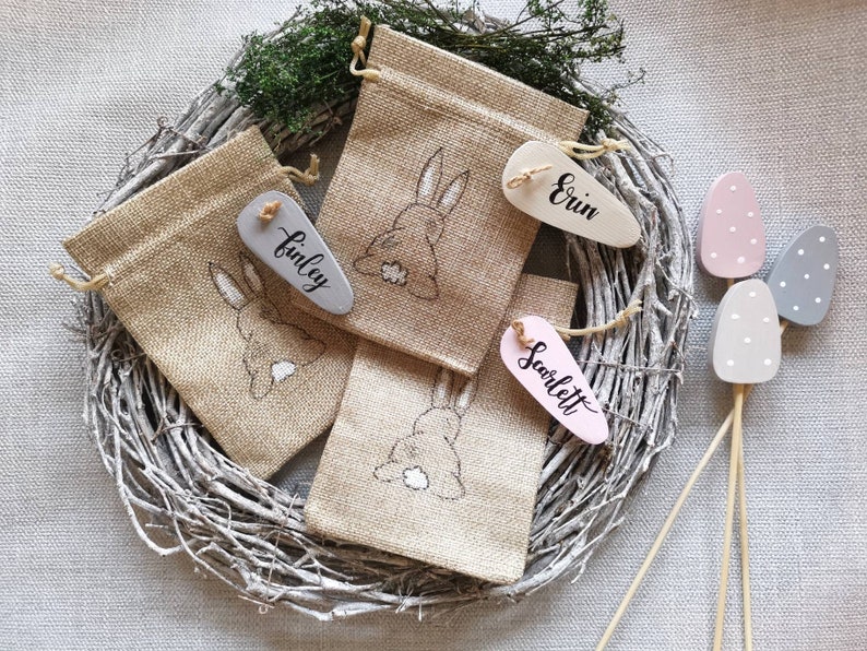 May include: Four small burlap bags with bunny embroidery, each with a wooden tag with a name written on it: Finley, Erin, Scarlett. The bags are arranged on a white twig wreath. Three wooden eggs with white polka dots are on sticks in the background.