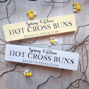 Op de afbeelding: Twee houten borden met de tekst "Spring Kitchen Hot Cross Buns Baked to Perfection" in zwarte letters op een witte achtergrond. Het bovenste bord is geel en het onderste bord is wit.