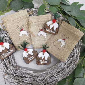 May include: Set of six Christmas-themed gift bags with personalized tags. The bags are made of burlap and feature a festive design of a Christmas pudding. The tags are wooden and have the names "Christina", "Alex", "Brian", and "Michael" written on them.