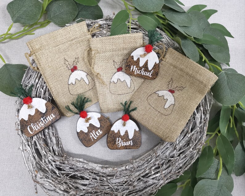 May include: Set of six Christmas-themed gift bags with personalized tags. The bags are made of burlap and feature a festive design of a Christmas pudding. The tags are wooden and have the names "Christina", "Alex", "Brian", and "Michael" written on them.