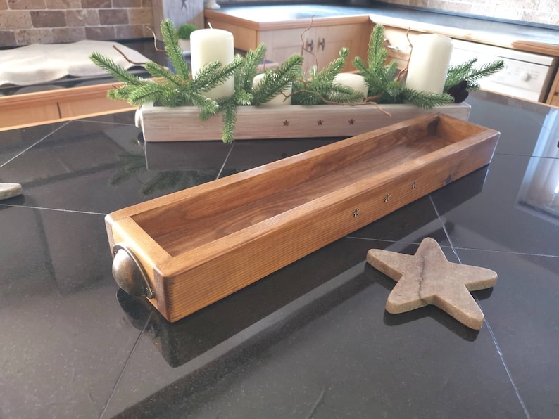Op de afbeelding: Een lange, rechthoekige houten plantenbak met een donkerbruine vlek. De plantenbak heeft metalen accenten op de hoeken en staat op een zwarte betegelde ondergrond.