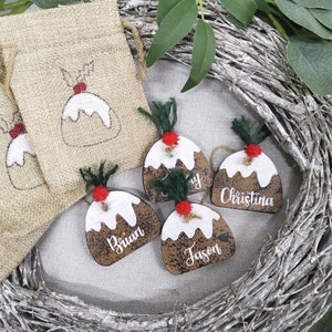 May include: Four wooden Christmas ornaments shaped like Christmas pudding with red and green accents. Each ornament has a different name written on it: Brian, Jason, Christina, and a partially visible name. The ornaments are displayed on a white background with a brown burlap bag and a twig wreath.