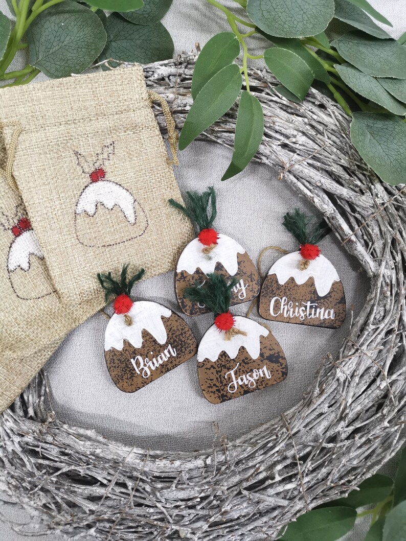 May include: Four wooden Christmas ornaments shaped like Christmas pudding with red and green accents. Each ornament has a different name written on it: Brian, Jason, Christina, and a partially visible name. The ornaments are displayed on a white background with a brown burlap bag and a twig wreath.