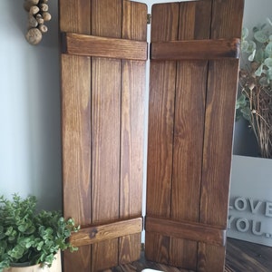 May include: Two brown wooden shutters with a brass hinge. The shutters are closed and have a rustic look. The shutters are on a wooden floor with a white chain link on the floor.