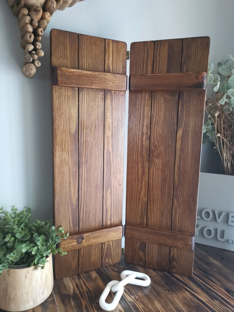 May include: Two brown wooden shutters with a brass hinge. The shutters are closed and have a rustic look. The shutters are on a wooden floor with a white chain link on the floor.