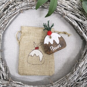 May include: A brown burlap gift bag with a white and red Christmas pudding design. A wooden Christmas pudding ornament with the name "Brian" is attached to the bag.