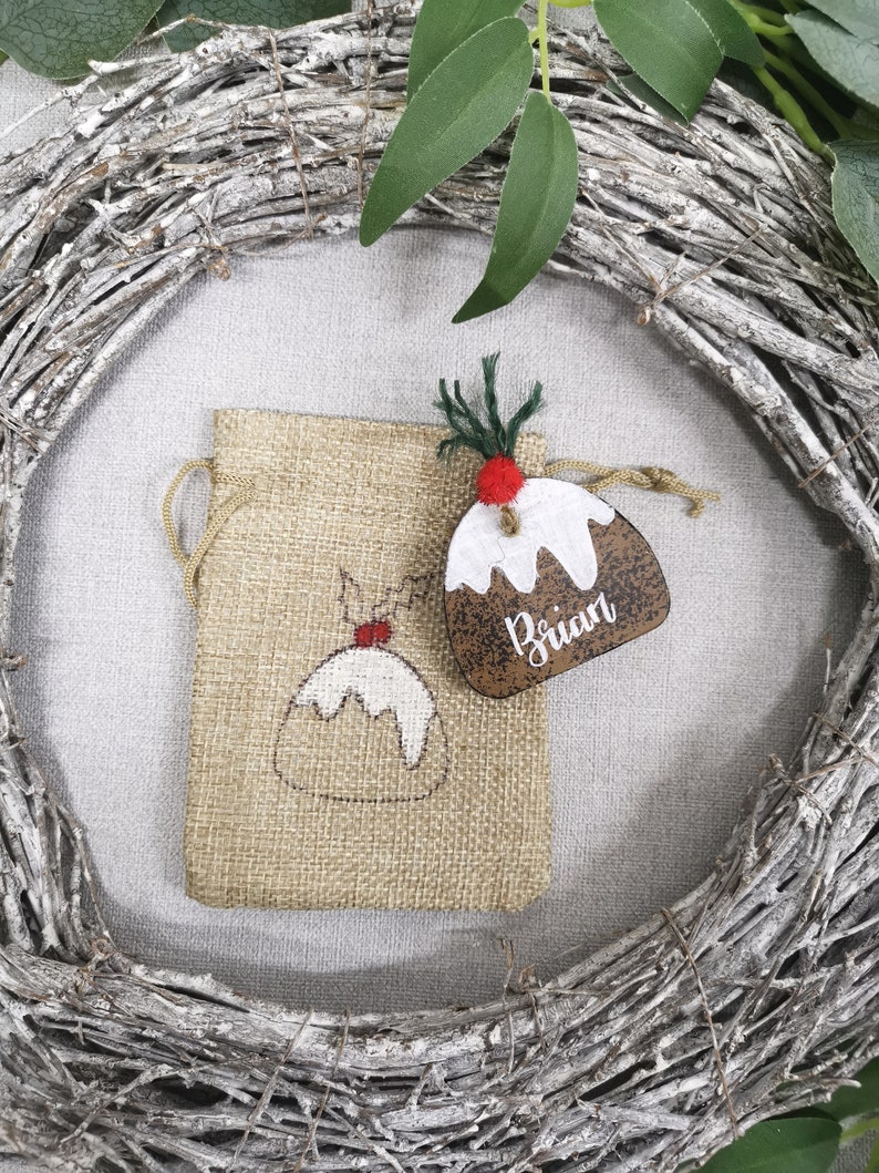 May include: A brown burlap gift bag with a white and red Christmas pudding design. A wooden Christmas pudding ornament with the name "Brian" is attached to the bag.