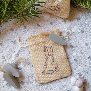 May include: Two small burlap bags with a bunny rabbit design. The bags are tied with twine and have a small wooden tag attached. The bags are sitting on a white background with small brown dots.
