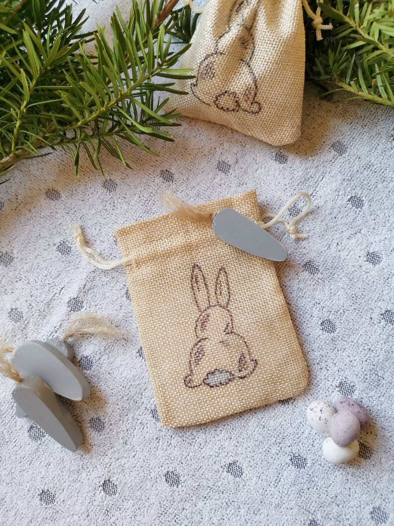 May include: Two small burlap bags with a bunny rabbit design. The bags are tied with twine and have a small wooden tag attached. The bags are sitting on a white background with small brown dots.