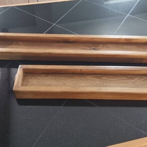 Op de afbeelding: Twee rechthoekige houten bakken, een groter dan de andere, beide met een natuurlijke houten afwerking. De bakken zijn leeg en staan op een zwarte betegelde ondergrond.