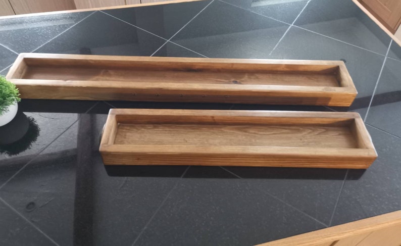 Op de afbeelding: Twee rechthoekige houten bakken, een groter dan de andere, beide met een natuurlijke houten afwerking. De bakken zijn leeg en staan op een zwarte betegelde ondergrond.