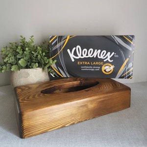 May include: A wooden tissue box holder with a dark brown stain. The box has a rectangular shape with a rounded oval opening in the center. A box of Kleenex Extra Large tissues is sitting on top of the holder.