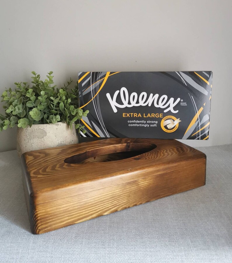 May include: A wooden tissue box holder with a dark brown stain. The box has a rectangular shape with a rounded oval opening in the center. A box of Kleenex Extra Large tissues is sitting on top of the holder.