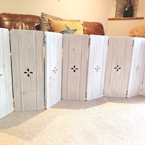 Op de afbeelding: Een witte, houten, opvouwbare huisdierpoort met een verweerde afwerking. Elk paneel heeft een decoratieve uitsparing en is verbonden door scharnieren. De poort is ontworpen om vrijstaand te zijn en is geschikt voor binnengebruik.