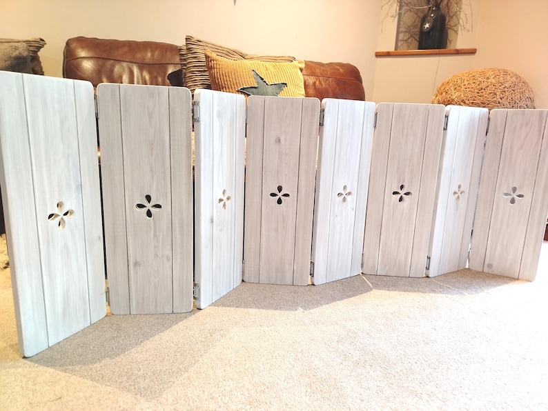 Op de afbeelding: Een witte, houten, opvouwbare huisdierpoort met een verweerde afwerking. Elk paneel heeft een decoratieve uitsparing en is verbonden door scharnieren. De poort is ontworpen om vrijstaand te zijn en is geschikt voor binnengebruik.