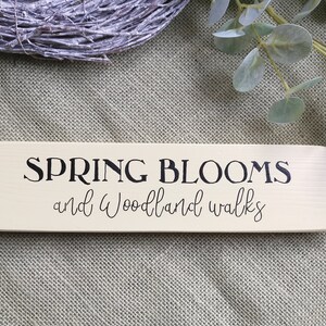 May include: A wooden sign with the text "SPRING BLOOMS and Woodland Walks" in black lettering on a cream background.