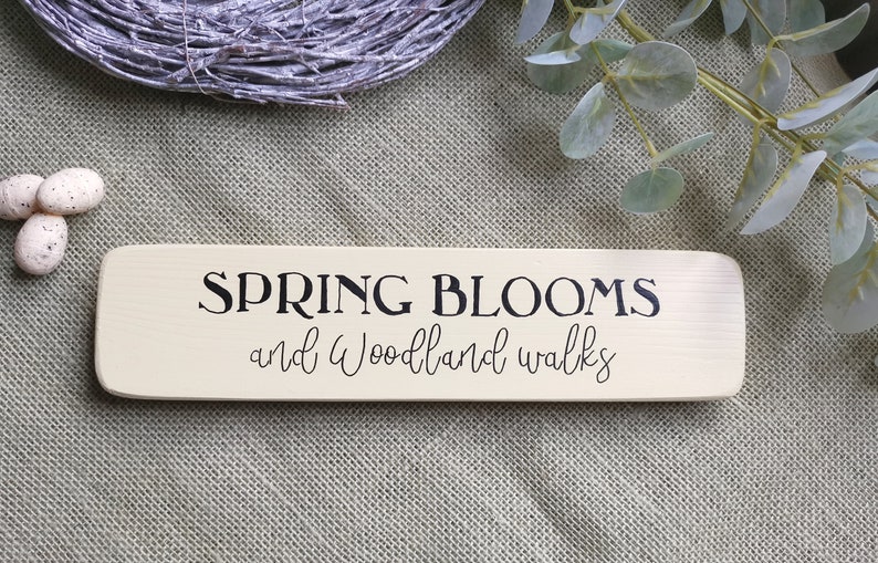 May include: A wooden sign with the text "SPRING BLOOMS and Woodland Walks" in black lettering on a cream background.