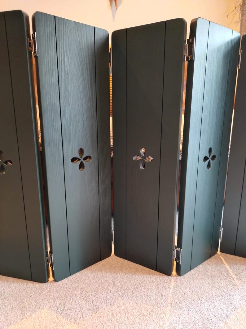 Op de afbeelding: Een donkergroen vouwscherm met vier panelen. Elk paneel heeft verticale houtnerf en een decoratieve vierbladige uitsparing. De panelen zijn verbonden door zilveren scharnieren.