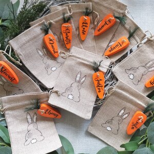 May include: Ten small burlap bags with orange carrot-shaped wooden tags. Each tag has a name written on it, including Stewart, David, Gareth, Eloise,  Joann,  Junia,  Austin,  Ebony,  Triston, and  Furley. The bags have a bunny rabbit design on them.