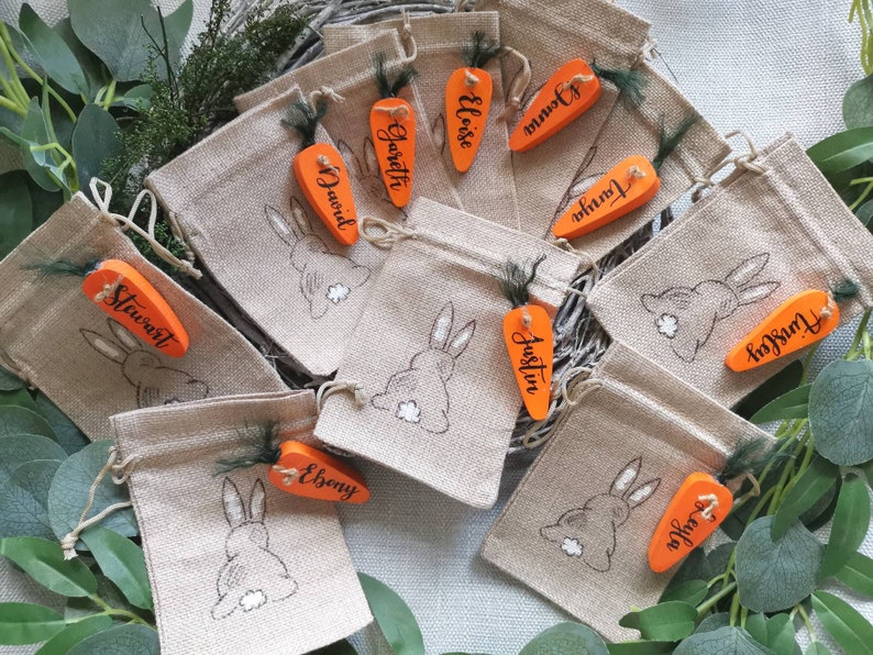 May include: Ten small burlap bags with orange carrot-shaped wooden tags. Each tag has a name written on it, including Stewart, David, Gareth, Eloise,  Joann,  Junia,  Austin,  Ebony,  Triston, and  Furley. The bags have a bunny rabbit design on them.