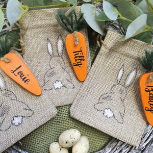 May include: Three burlap gift bags with bunny designs and carrot-shaped tags. The tags are orange and have the names "Louie", "Tilly", and "Harvey" written on them. The bags are sitting on a woven wreath with three speckled eggs.