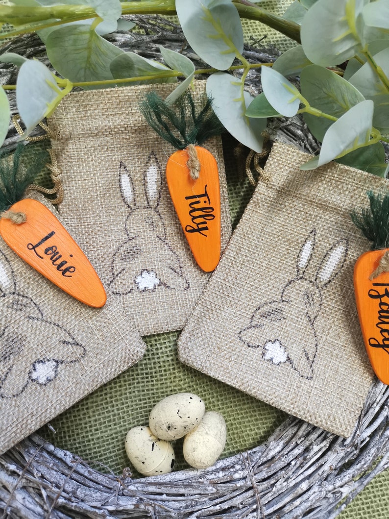 May include: Three burlap gift bags with bunny designs and carrot-shaped tags. The tags are orange and have the names "Louie", "Tilly", and "Harvey" written on them. The bags are sitting on a woven wreath with three speckled eggs.