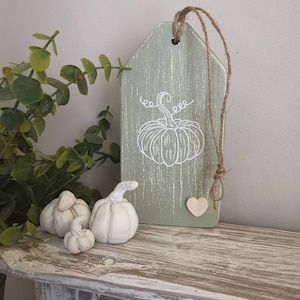 May include: A green wooden tag with a white pumpkin design and a twine hanger. The tag has a distressed finish and a small heart-shaped wooden embellishment. Three small white ceramic pumpkins are in the foreground.