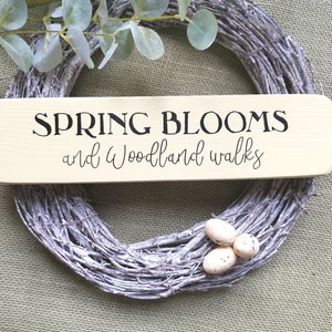 May include: A wooden sign with the text "SPRING BLOOMS and woodland walks" in black lettering on a light beige background. The sign is sitting on top of a white twig wreath.