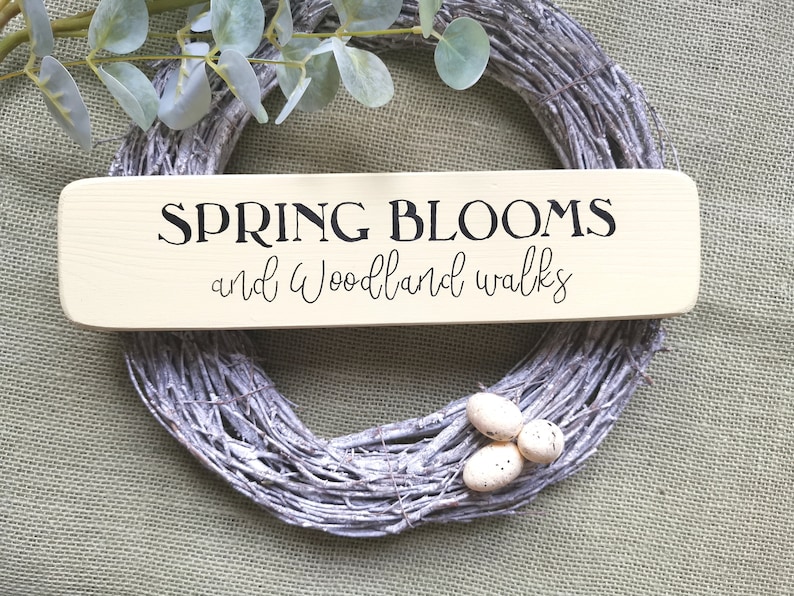 May include: A wooden sign with the text "SPRING BLOOMS and woodland walks" in black lettering on a light beige background. The sign is sitting on top of a white twig wreath.