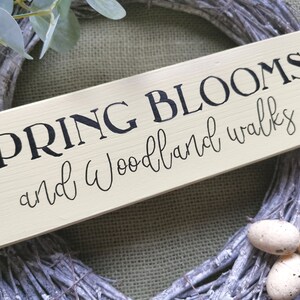 May include: A light yellow wooden sign with black text that reads "SPRING BLOOMS and woodland walks". The sign is leaning against a gray and white twig wreath.