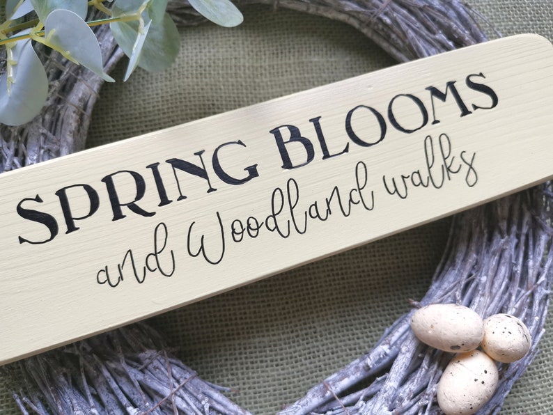 May include: A light yellow wooden sign with black text that reads "SPRING BLOOMS and woodland walks". The sign is leaning against a gray and white twig wreath.