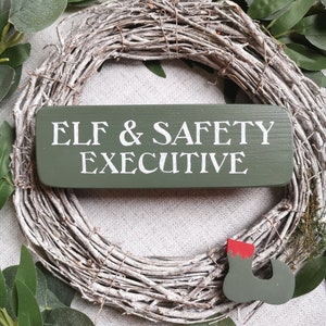 May include: A green wooden sign with white text that reads "Elf & Safety Executive". The sign is hanging on a white twig wreath with green leaves.