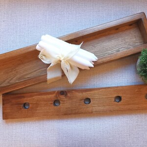 Op de afbeelding: Een houten dienblad met een rechthoekige vorm en een kleiner houten dienblad met vijf gaten. Het grotere dienblad heeft een stapel witte kaarsen die met een lint zijn vastgebonden.