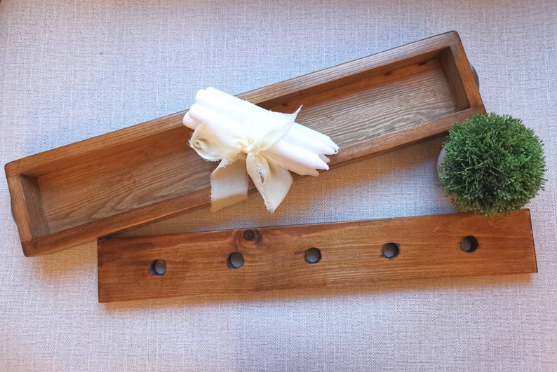 Op de afbeelding: Een houten dienblad met een rechthoekige vorm en een kleiner houten dienblad met vijf gaten. Het grotere dienblad heeft een stapel witte kaarsen die met een lint zijn vastgebonden.