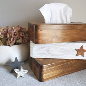 May include: Three wooden tissue box covers stacked on top of each other. The top cover is a dark brown wood, the middle cover is white with a distressed look, and the bottom cover is a dark brown wood. The middle cover has a rusty star on the front.