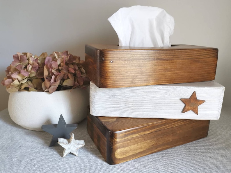 May include: Three wooden tissue box covers stacked on top of each other. The top cover is a dark brown wood, the middle cover is white with a distressed look, and the bottom cover is a dark brown wood. The middle cover has a rusty star on the front.